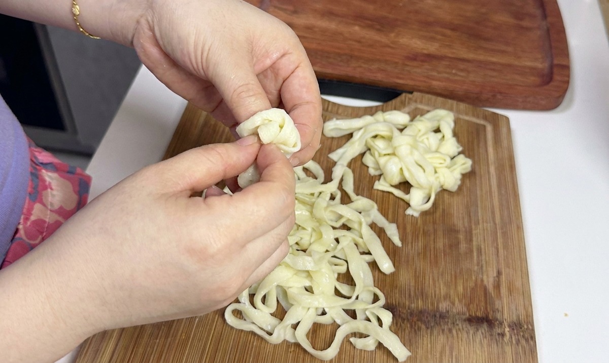 Manos separando y soltando cuidadosamente una pila de fideos fríos blancos, anchos y planos sobre una tabla de cortar de madera.