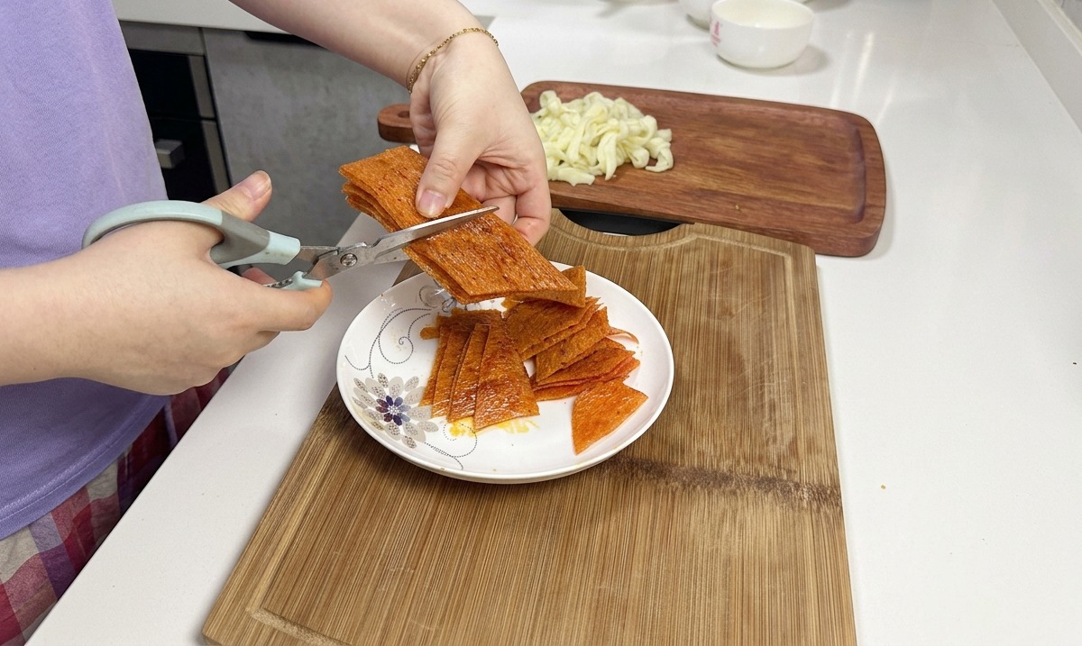 Tijeras de cocina cortando una pila de tiras naranjas picantes de latiao sobre un plato de cerámica blanca.