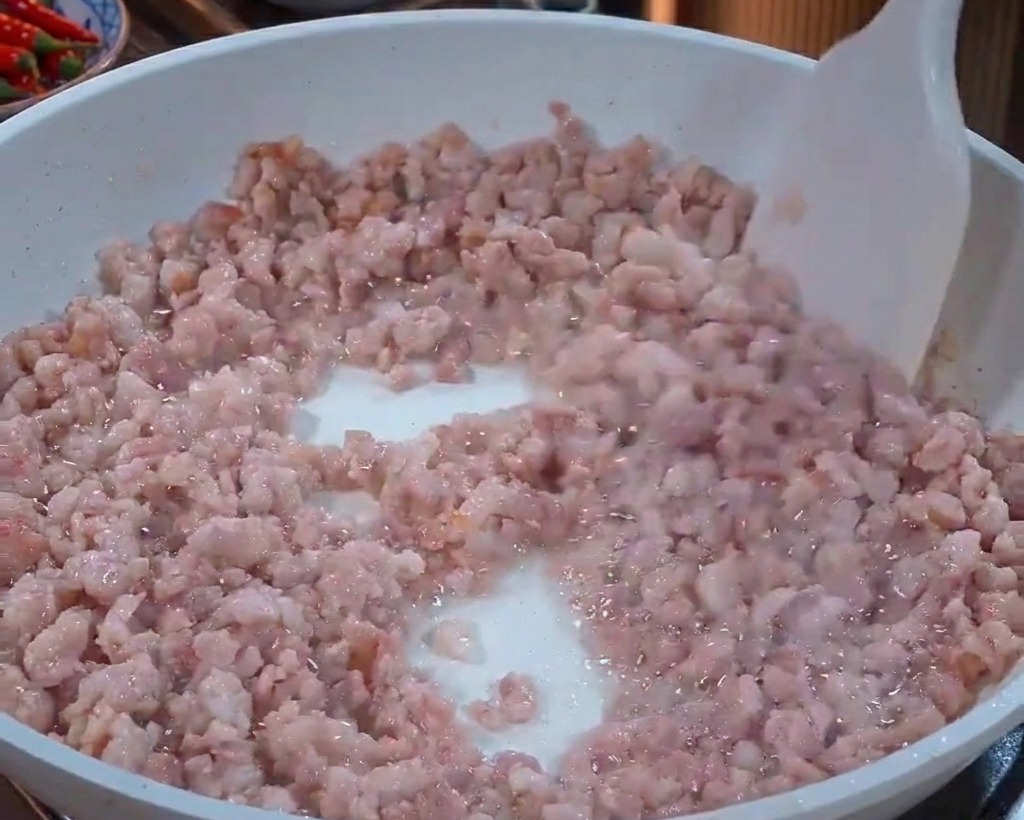 Carne de cerdo picada removiéndose en una sartén blanca con una espátula blanca a medida que cambia de color rosa a gris claro.