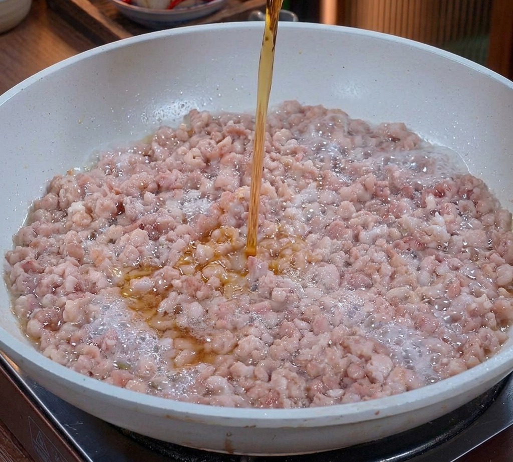 Un líquido ámbar claro vertiéndose desde un vaso sobre la carne de cerdo picada que se saltea en una sartén.