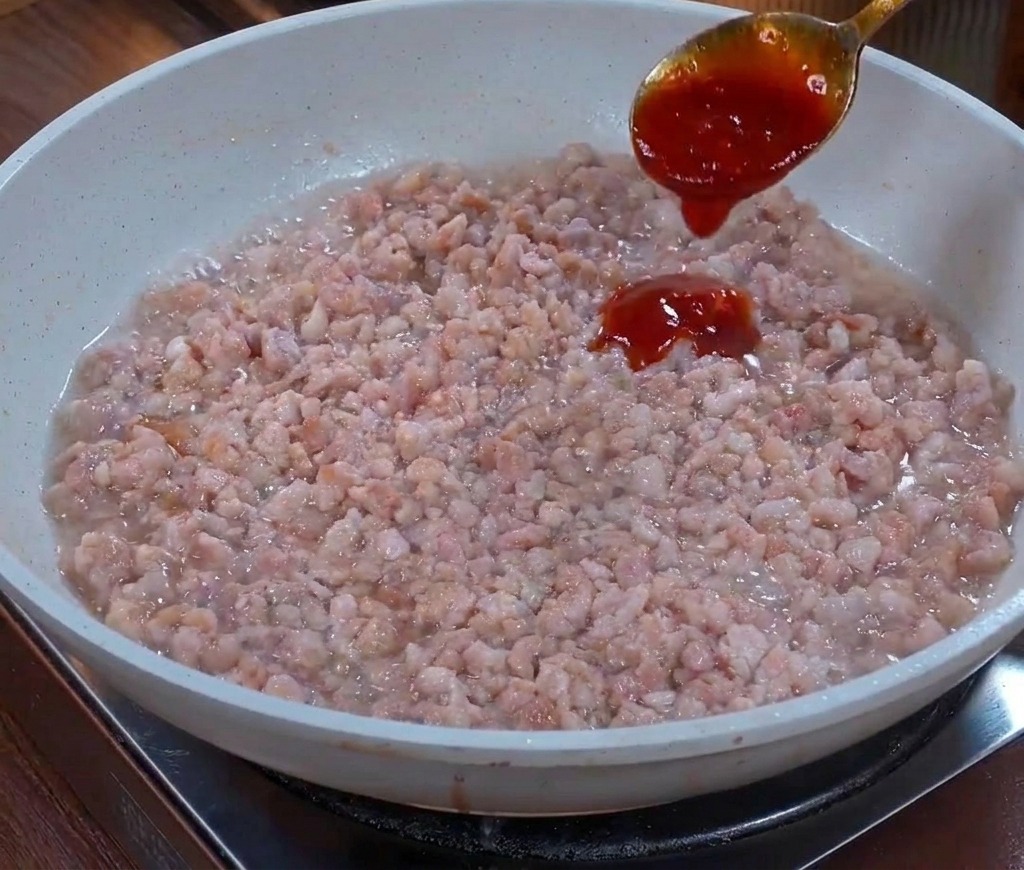 Una cuchara de metal vertiendo una salsa espesa de color marrón oscuro sobre una base de carne picada salteada en una sartén.