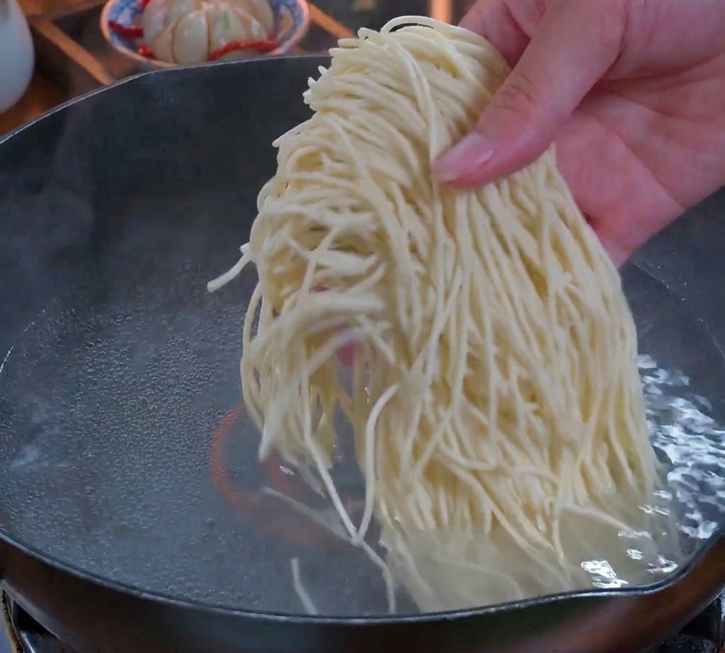 Una mano soltando un fajo espeso de fideos finos y frescos en una olla con agua hirviendo.