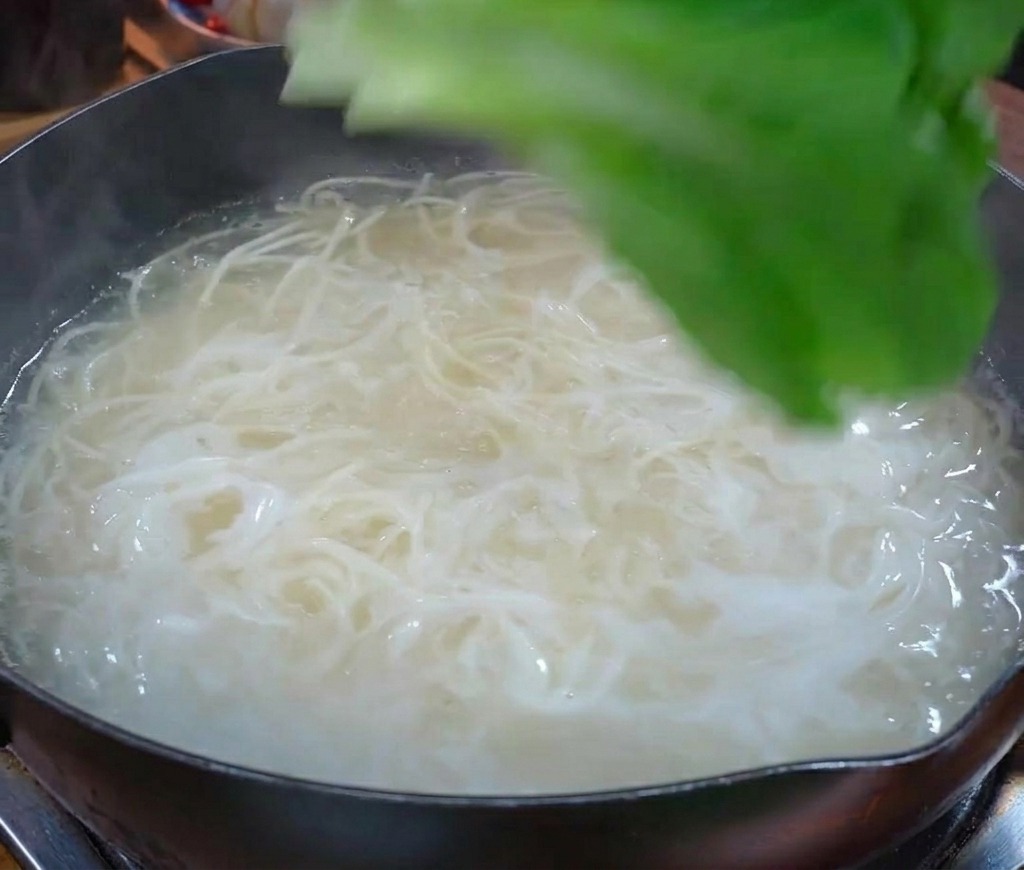 Vegetales frescos de hoja verde añadiéndose a una olla de agua hirviendo que contiene fideos blancos.