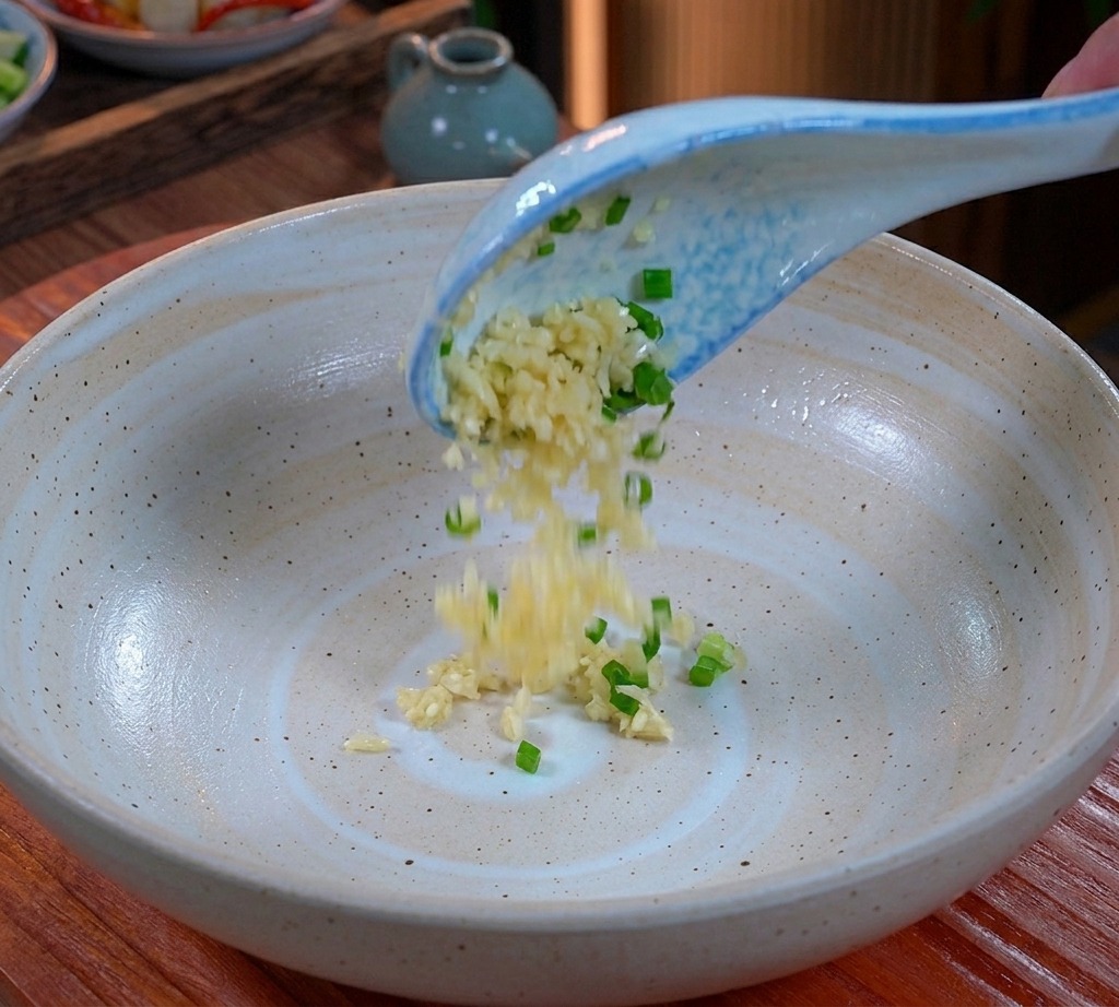 Ajo picado y cebolleta picada cayendo desde una cuchara azul y blanca al fondo de un tazón de cerámica ancho.