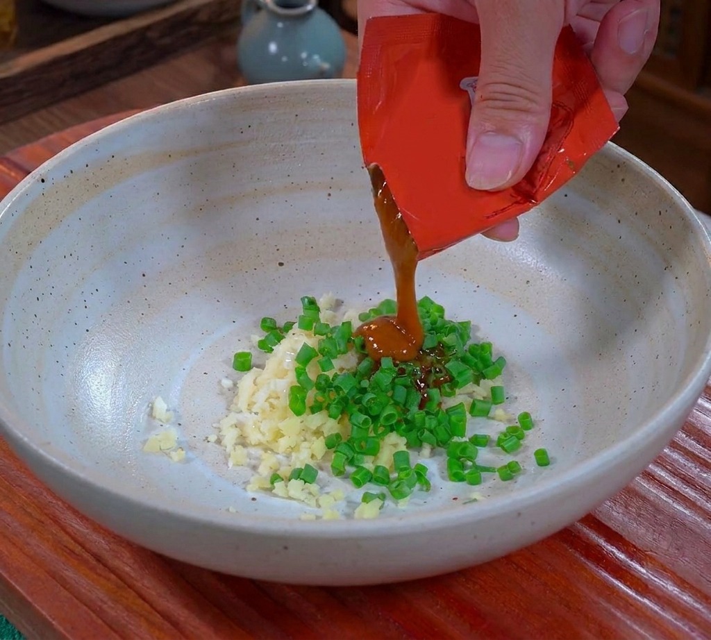 Una mano exprimiendo salsa Dandan espesa y marrón de un paquete rojo en un tazón de cerámica sobre ajo y cebolletas picados.
