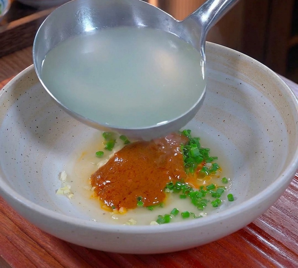 Agua caliente y turbia de la cocción de los fideos vertiéndose con un cucharón en un tazón que contiene condimentos picantes y grasa de cerdo.