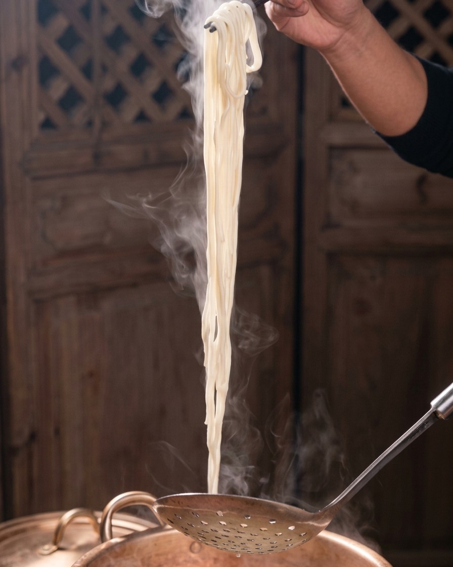 Un buen puñado de fideos blancos humeantes sacándose de una olla de cobre con unos palillos oscuros y una espumadera metálica.