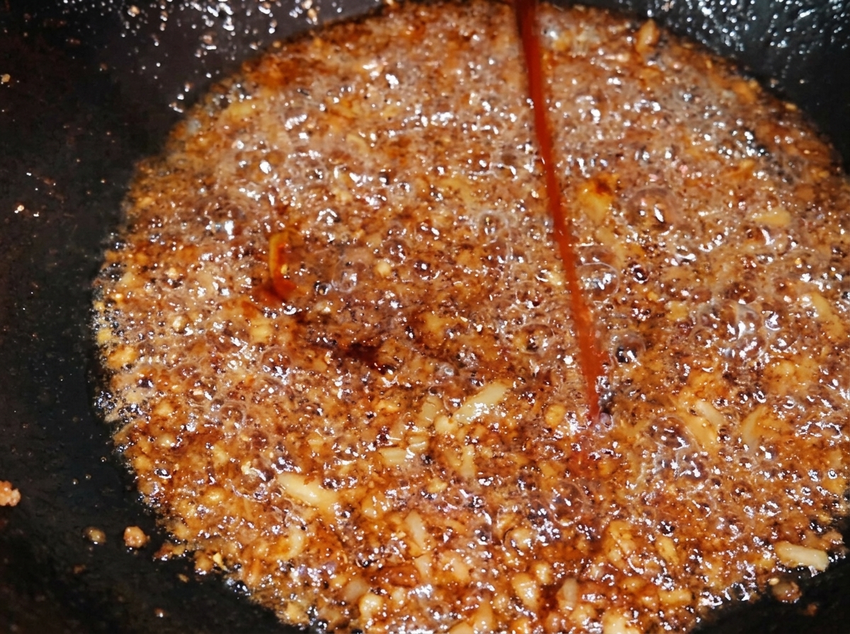 Chorrito de condimento de color caramelo oscuro cayendo sobre el cerdo sazonado en un wok, intensificando su tono.