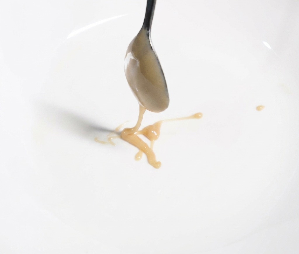 Cuchara de metal echando pasta de sésamo espesa de color marrón claro en un bol blanco.