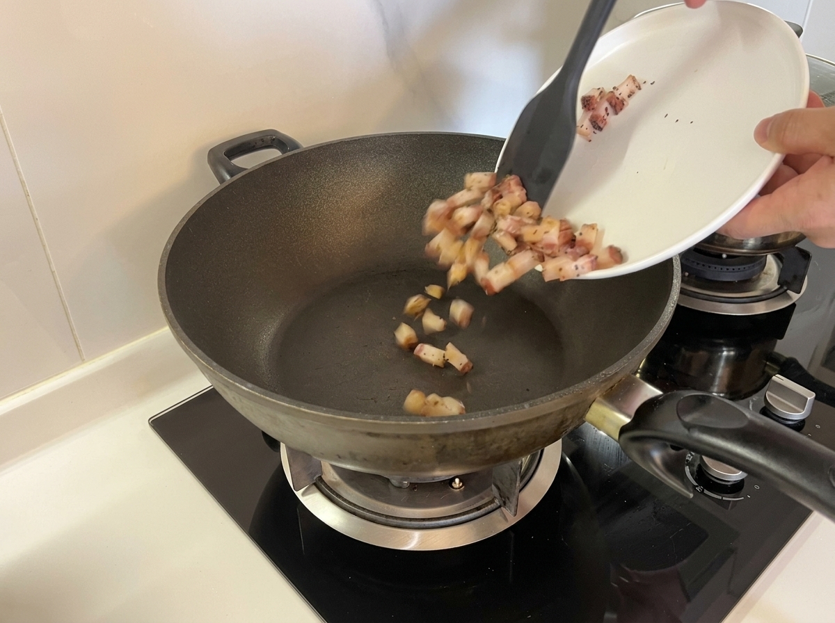 Dados de guanciale crudo vertidos desde un plato blanco a un wok seco sin calentar usando una espátula.