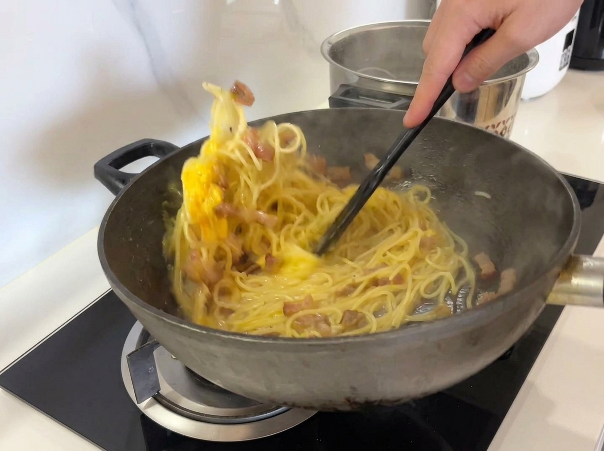 Removiendo enérgicamente espaguetis, guanciale y la mezcla de huevo amarillo en un wok con una espátula negra.