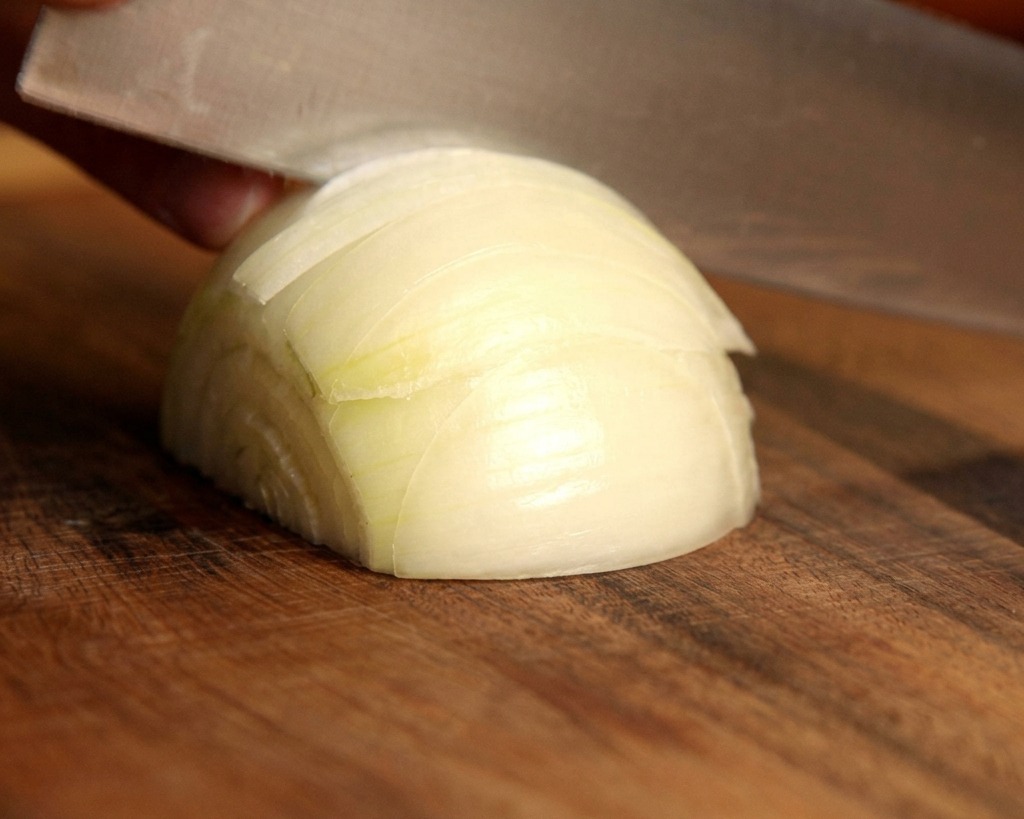 Un cuchillo afilado cortando una cebolla blanca fresca en plumas finas sobre una tabla de cortar de madera.