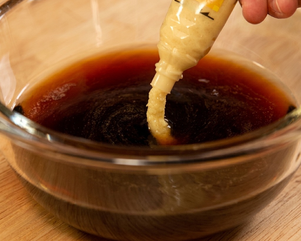 Pasta de jengibre rallado exprimiéndose desde un tubo hacia una mezcla de salsa oscura a base de soja en un tazón de vidrio.
