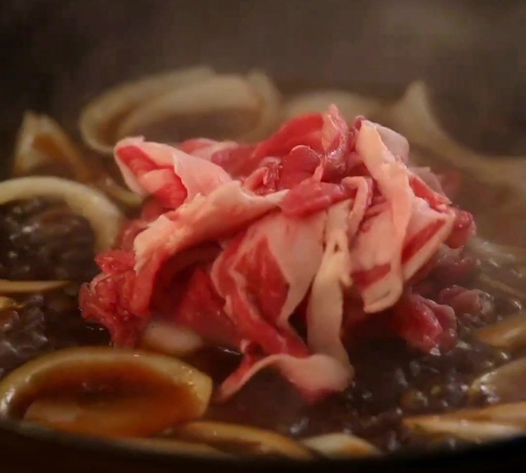 Láminas de panceta de res cruda añadiéndose a una sartén con cebollas y salsa japonesa en ebullición.