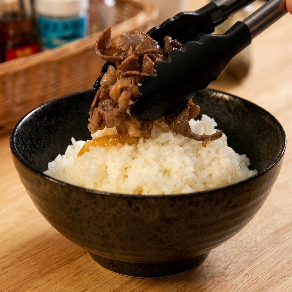 Pinzas negras colocando con cuidado una porción generosa de carne y cebolla cocinadas sobre un tazón de arroz blanco.