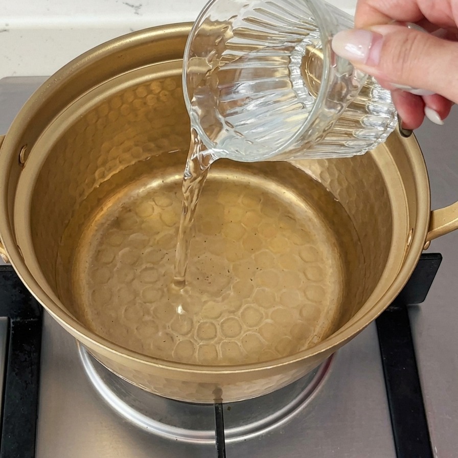 Agua fría vertiéndose desde un vaso de cristal en una pequeña olla de metal dorado sobre una estufa de gas.