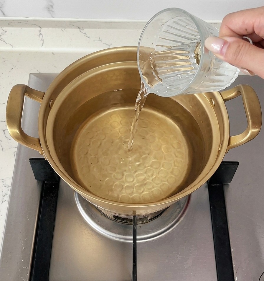 Una mano vertiendo una taza de agua fría en una olla dorada que ya contiene agua caliente sobre la estufa.