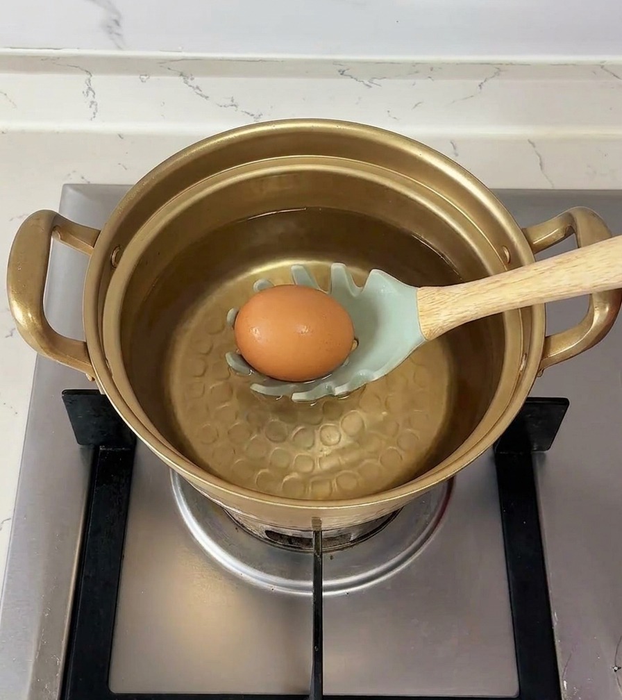 Un huevo marrón apoyado en una cuchara con ranuras de color verde claro que se sumerge con cuidado en una olla con agua.