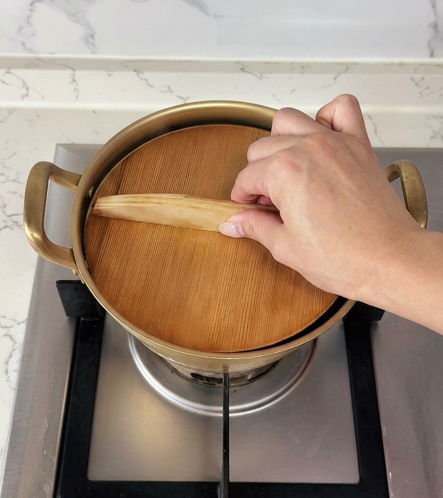 Una mano colocando una tapa de madera plana sobre una pequeña olla dorada para cubrirla.