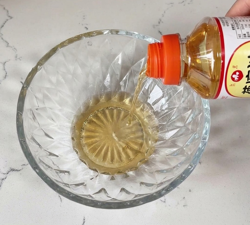 Líquido transparente de mirin vertiéndose desde una botella en un bol de cristal sobre una encimera blanca.