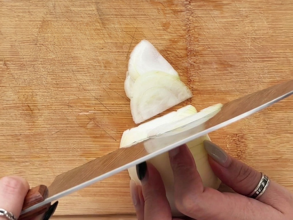 Manos cortando una cebolla blanca en finas plumas sobre una tabla de cortar de madera.
