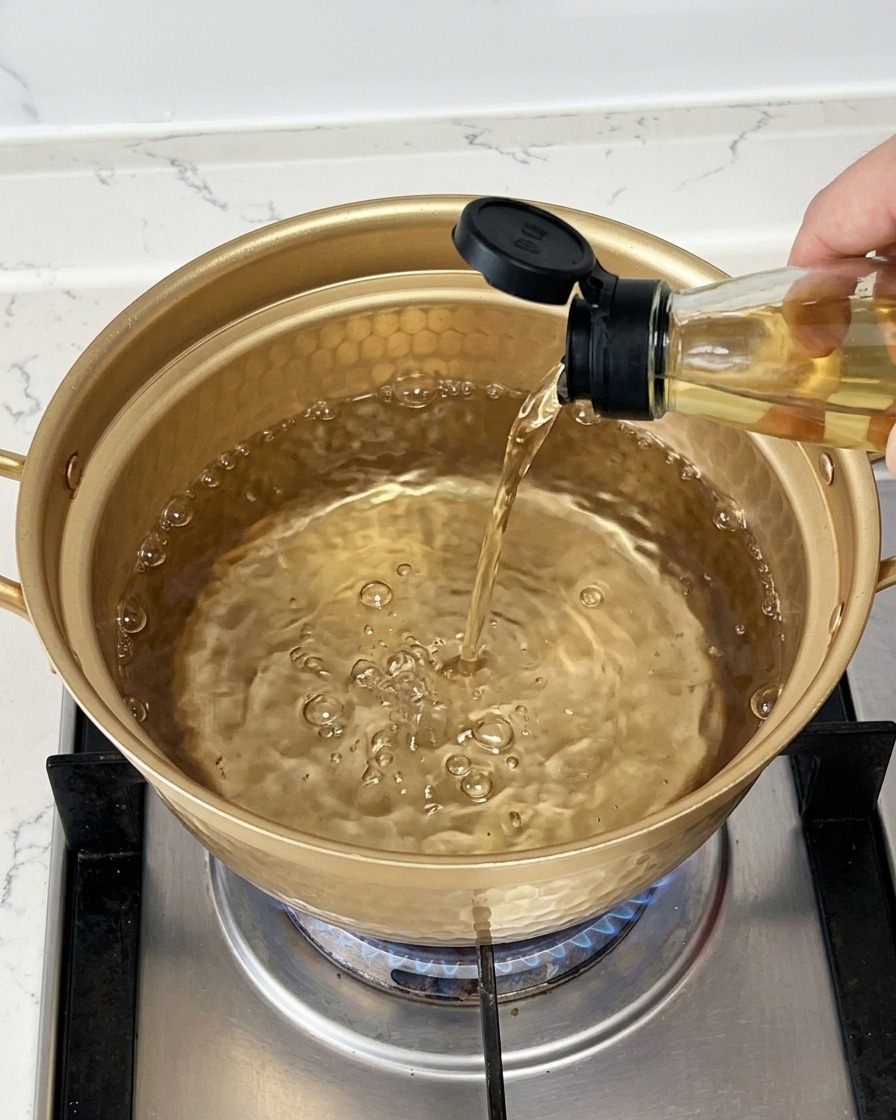 Vertiendo vino de cocina transparente desde una botella de vidrio en una olla dorada llena de agua hirviendo.