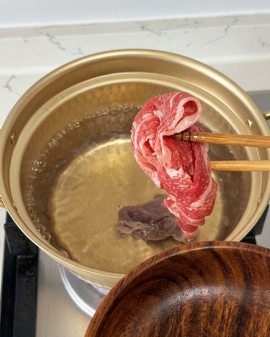 Sosteniendo una fina lámina de ternera cruda y veteada con palillos sobre una olla dorada de agua hirviendo.