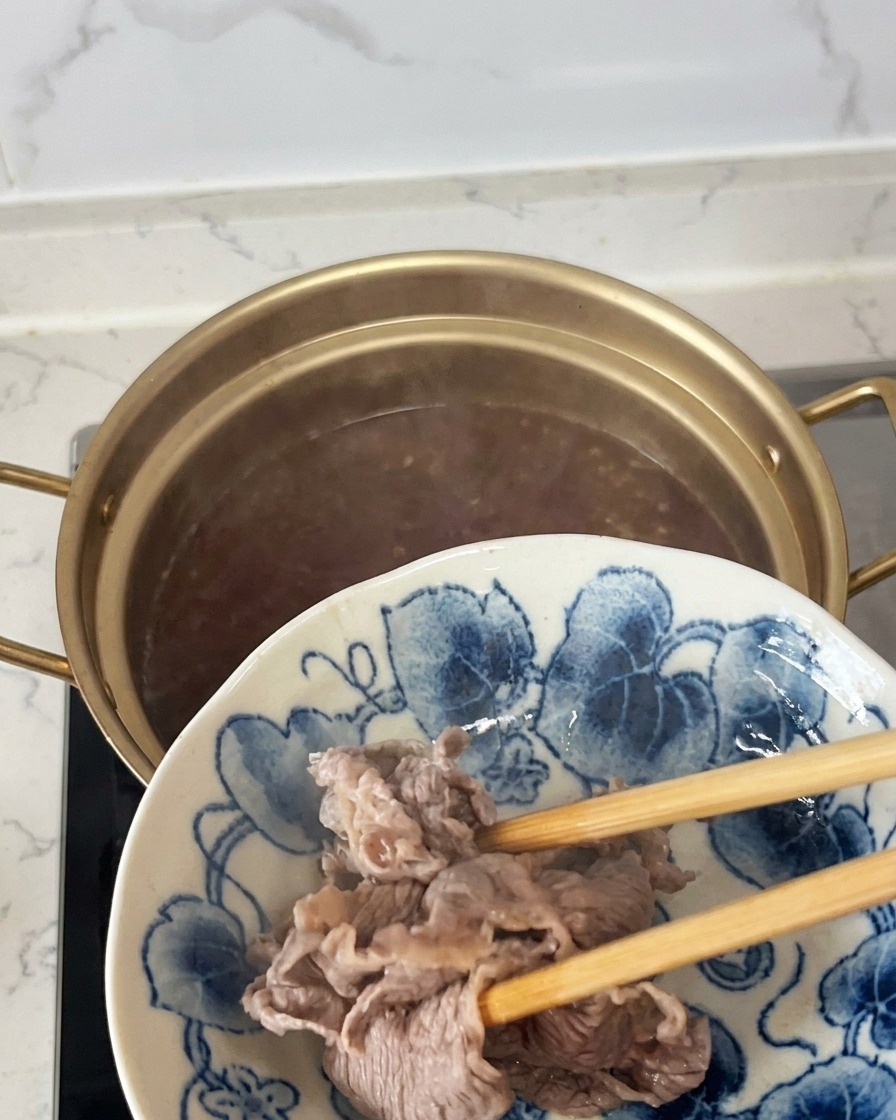 Retirando láminas de ternera semi-cocinadas y opacas de una olla con palillos y pasándolas a un plato con motivos azules y blancos.
