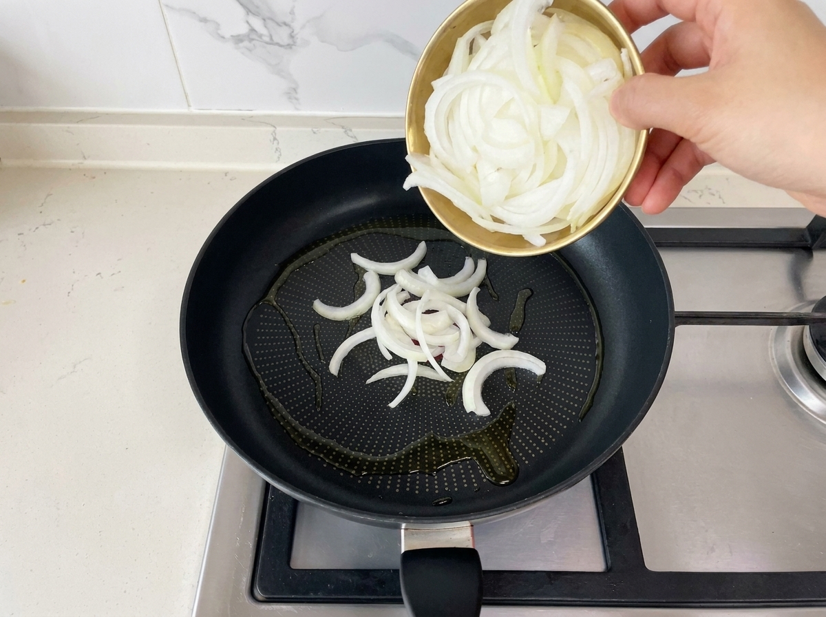 Vertiendo cebollas blancas recién cortadas desde un pequeño bol dorado a una sartén negra con aceite caliente.