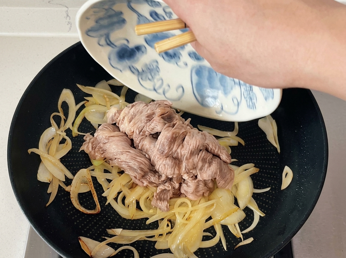 Palillos deslizando láminas de ternera escaldadas desde un plato decorado hacia una sartén con cebollas sofritas.