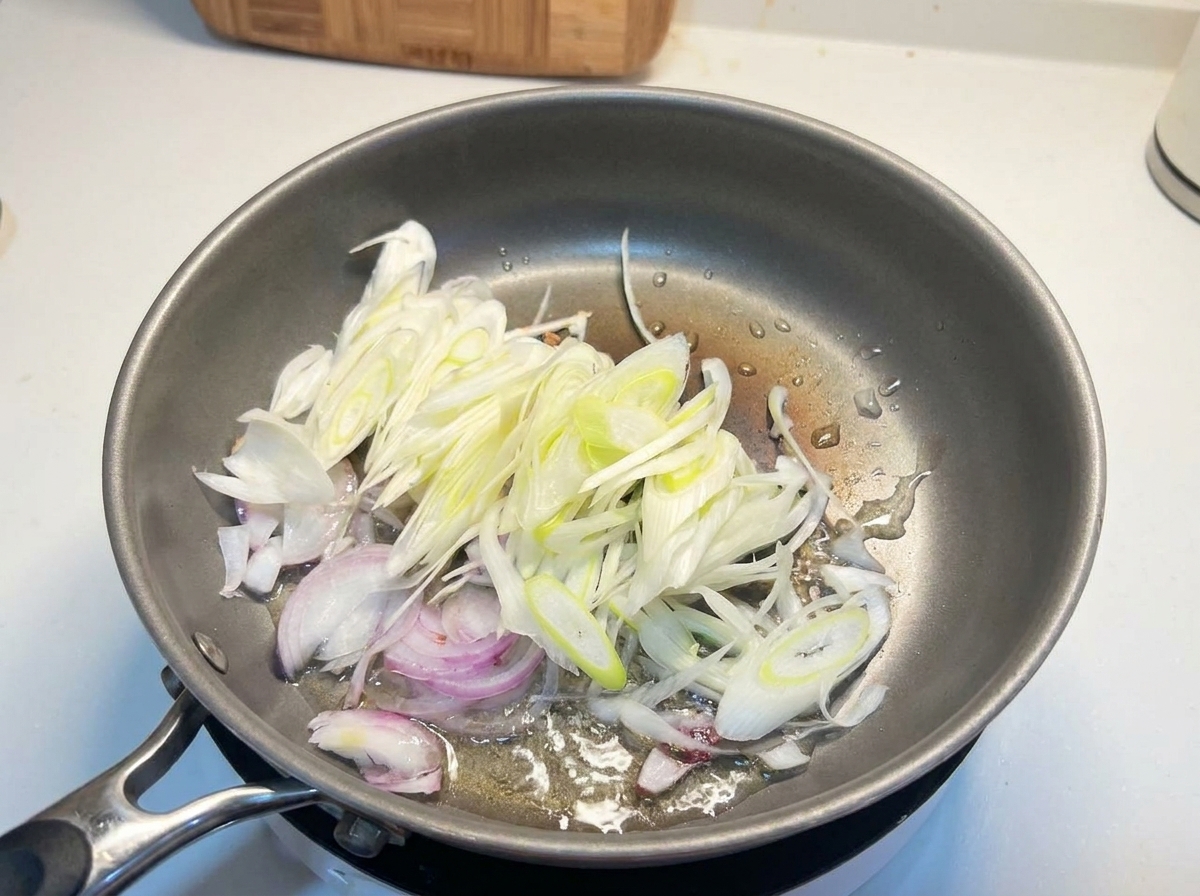 Láminas de cebolla blanca y cebolleta chisporroteando en aceite caliente dentro de una sartén de metal.