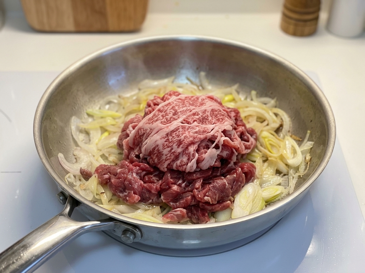 Un montón de carne de ternera cruda, con grasa y cortada muy fina, reposando sobre cebollas cocinadas en una sartén de metal.
