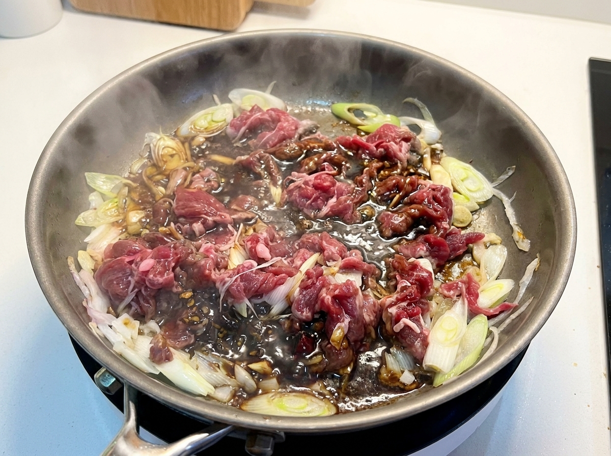 Ternera y cebollas parcialmente cocinadas en una sartén con una salsa oscura, rica y burbujeante.