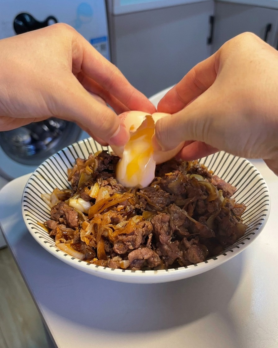 Dos manos abriendo un huevo onsen, dejando caer una yema suave y una clara semilíquida sobre un tazón humeante de arroz con ternera.