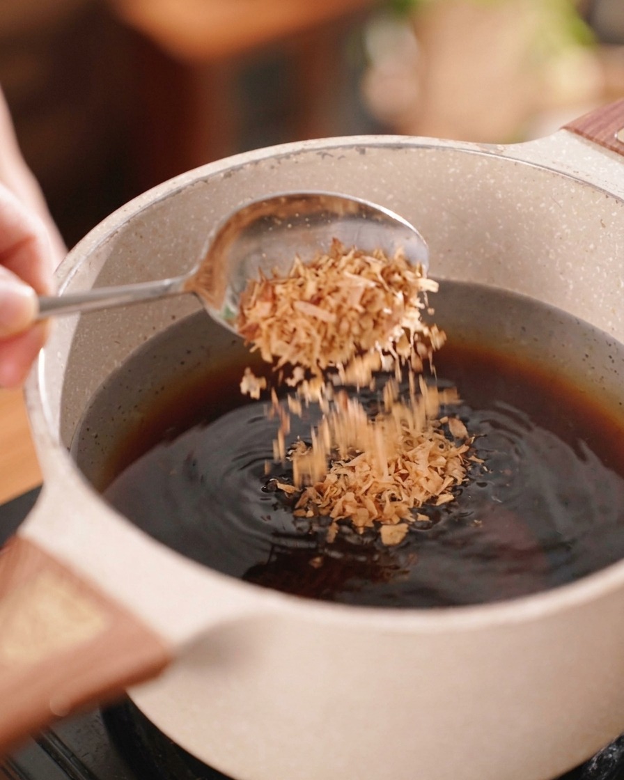 Una persona usando una cuchara de plata para espolvorear chalotas fritas de color marrón crujiente en una olla de marinado oscuro.