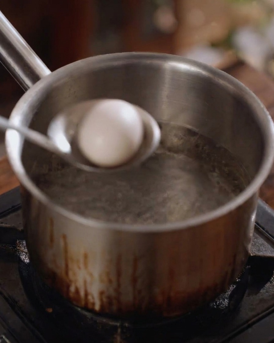 Un huevo blanco entero siendo bajado suavemente a una olla de agua hirviendo con un cucharón de metal.