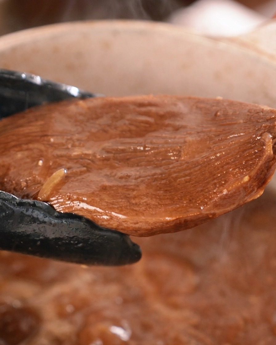 Un trozo grueso de lengua de res estofada siendo levantado de una olla de salsa oscura con pinzas de cocina negras.