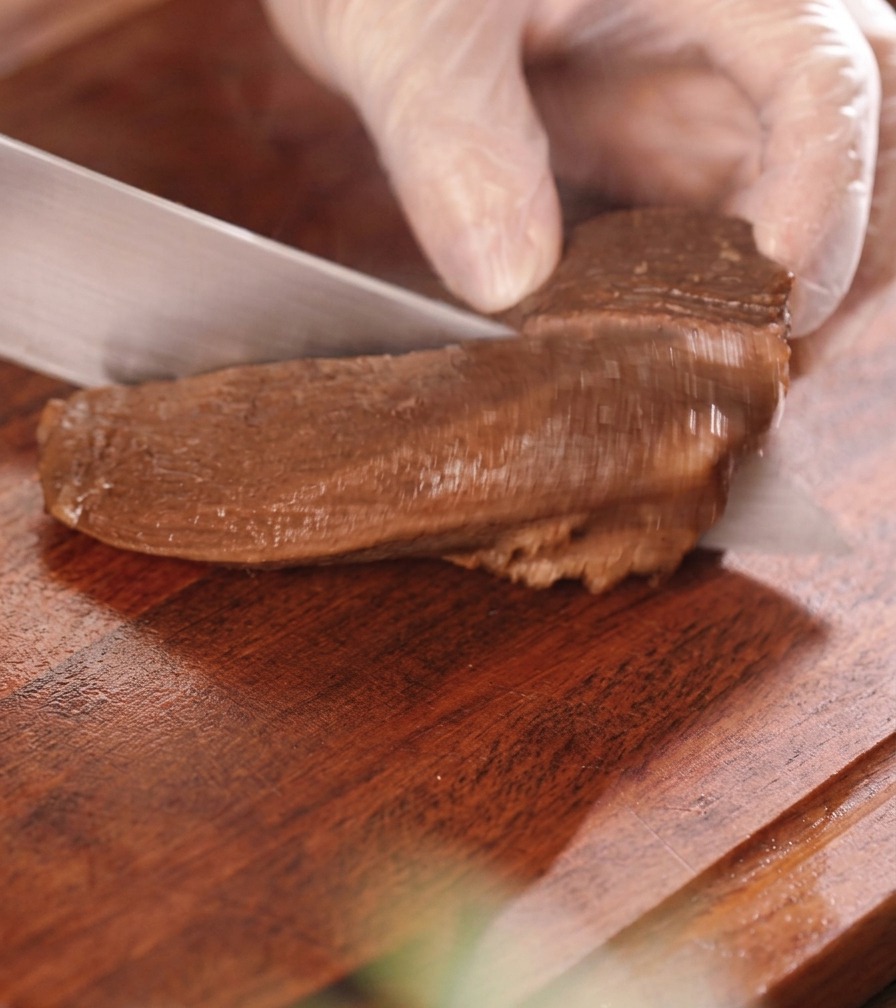 Una mano con guante cortando un trozo de lengua de res estofada y oscura en trozos gruesos de tamaño de un bocado sobre una tabla de madera.
