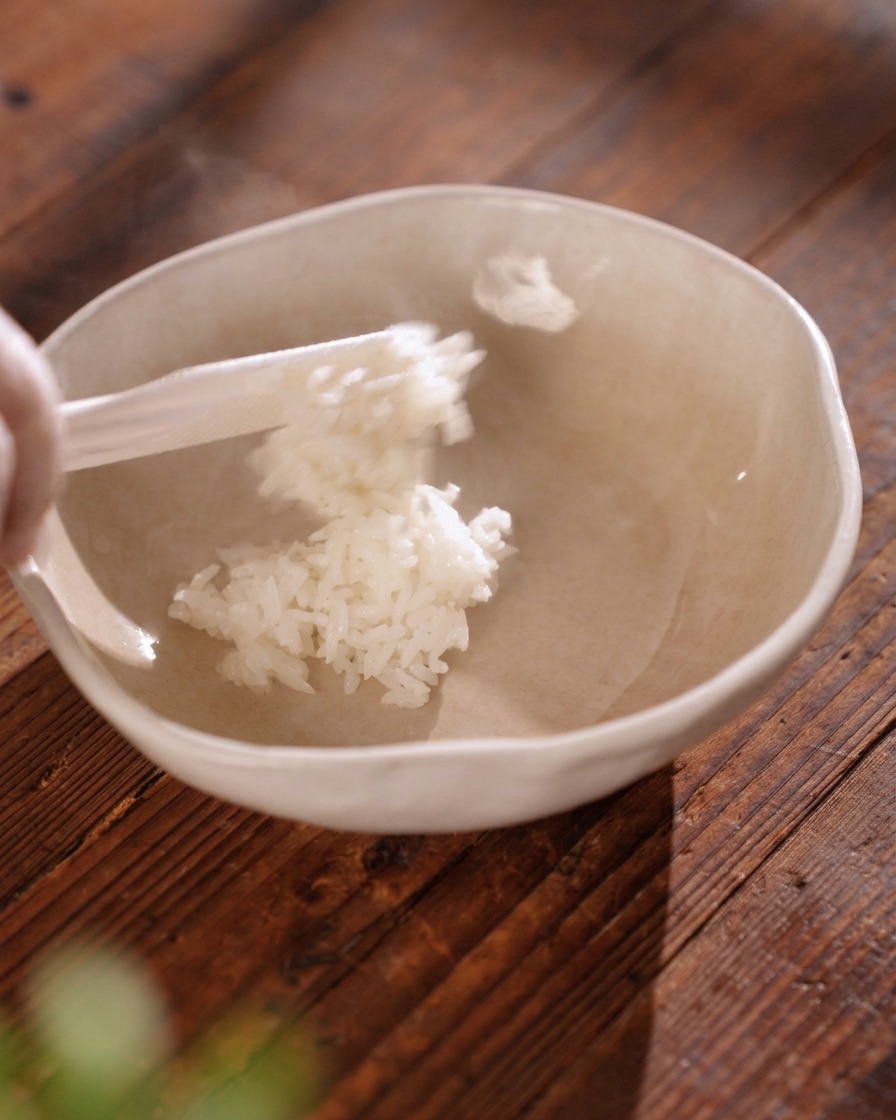 Una paleta de arroz transfiriendo arroz blanco esponjoso y humeante a un tazón de cerámica beige claro.