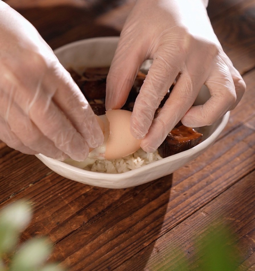 Manos con guantes rompiendo con cuidado una cáscara de huevo blanda para soltar un huevo escalfado en un tazón de arroz con carne.