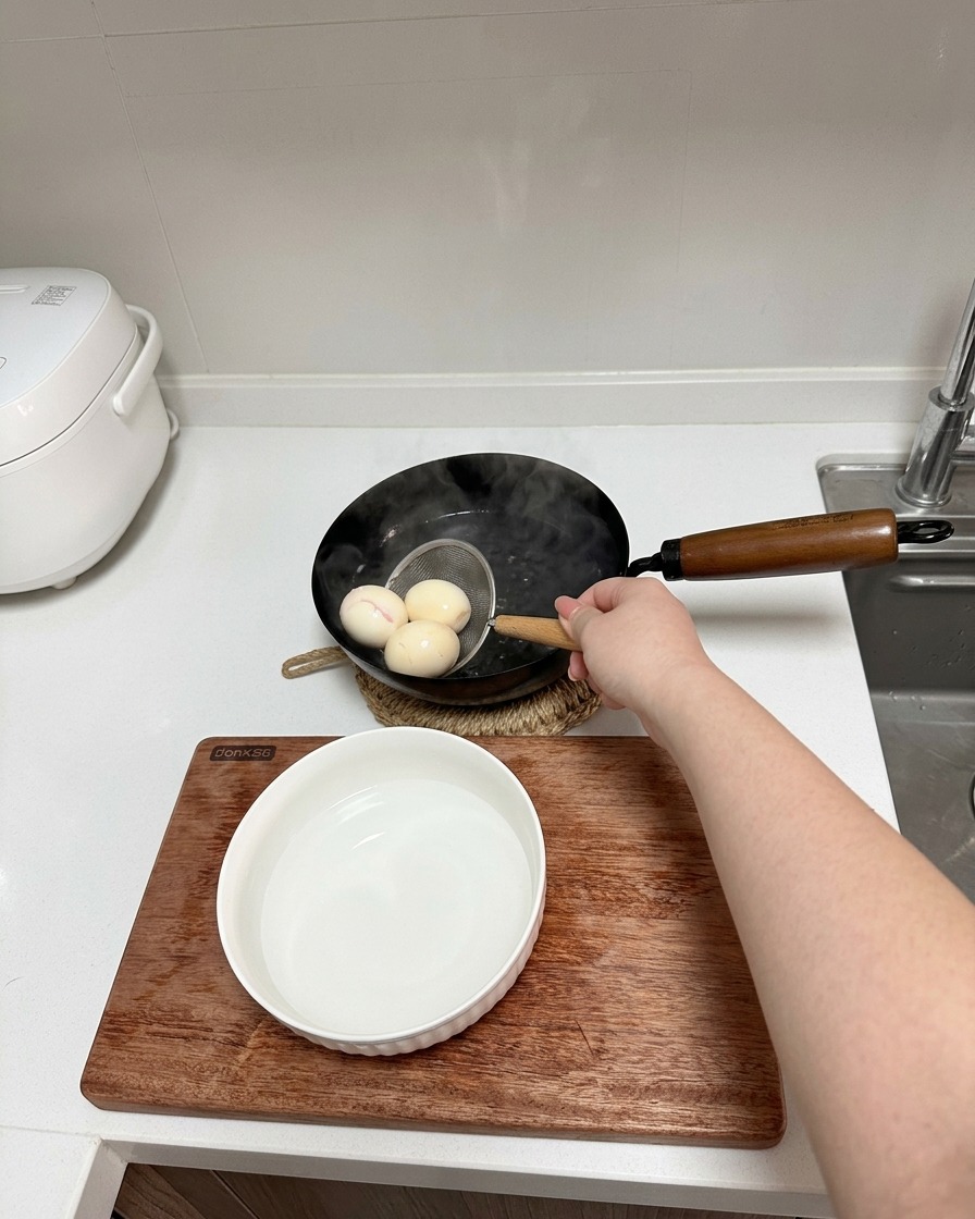 Una mano usando un pequeño colador de alambre para sacar tres huevos cocidos de una olla hacia un tazón con agua fría.