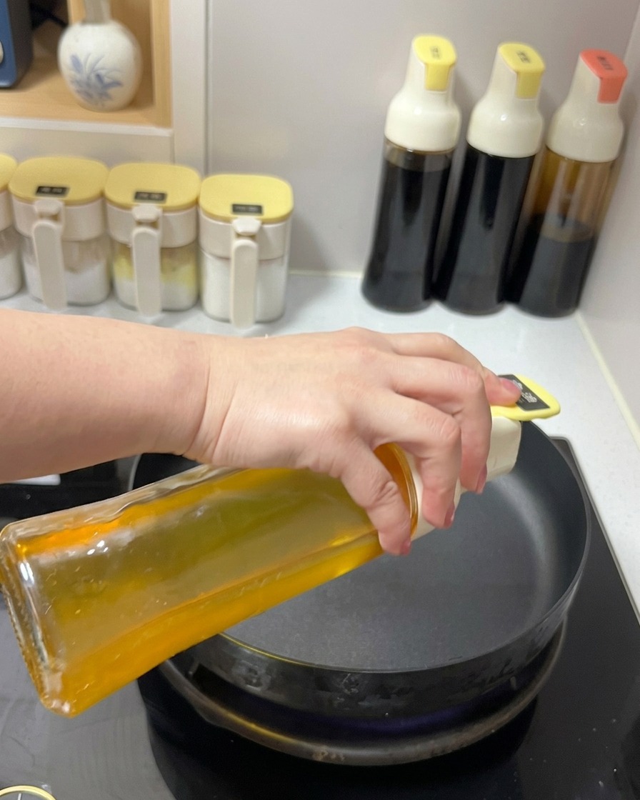 Aceite de cocina transparente siendo vertido desde un dispensador de vidrio en una sartén negra sobre la estufa.