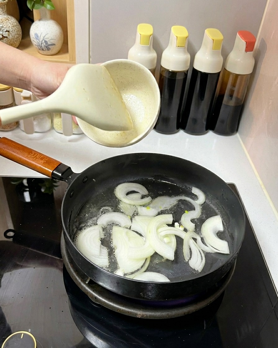 Tiras de cebolla blanca siendo removidas en una sartén con aceite usando una espátula blanca.