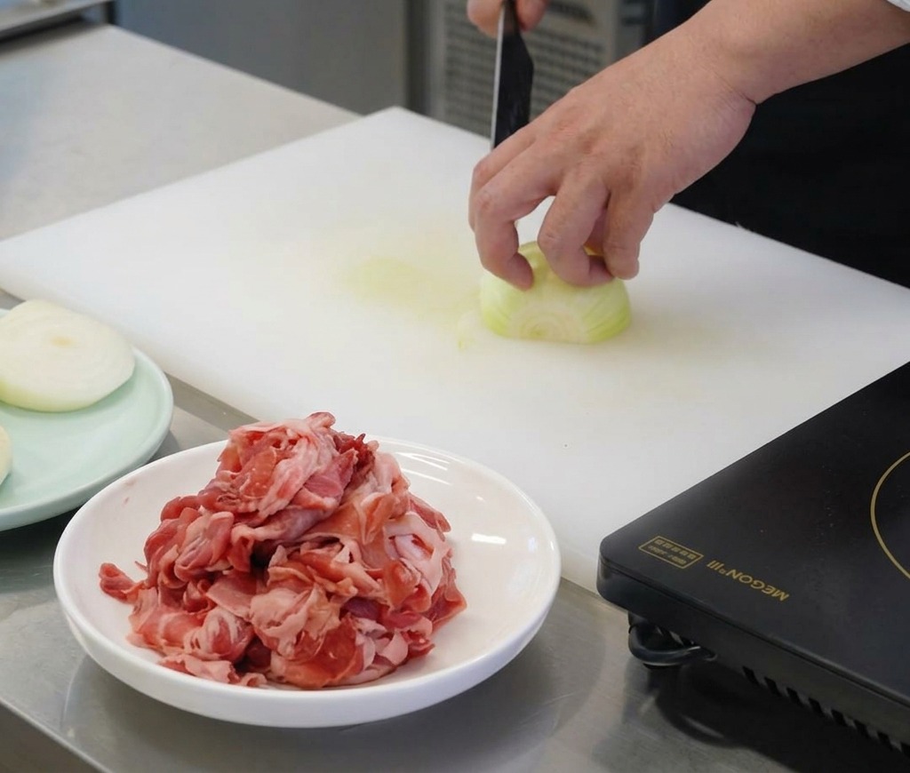 La mano de un chef sosteniendo una cebolla blanca sobre una tabla de cortar blanca mientras la corta en tiras finas con un cuchillo afilado.