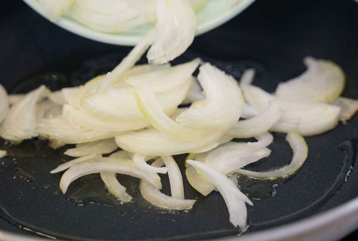 Tiras de cebolla blanca cruda siendo vertidas desde un plato verde claro en una sartén negra con aceite de cocina brillante.