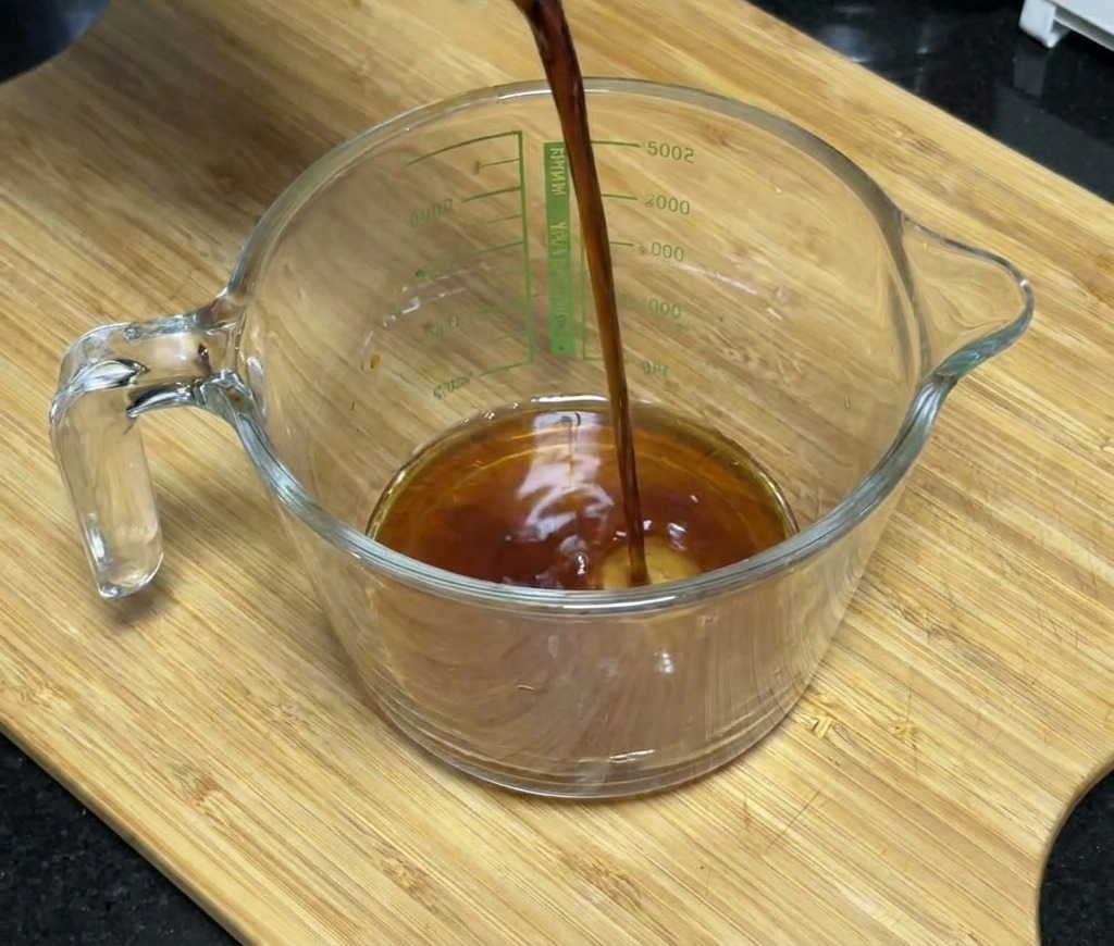 Salsa de soja oscura vertiéndose en una taza medidora de cristal que contiene miel y condimentos.