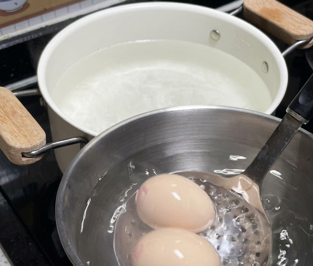 Dos huevos reposando en un tazón de acero inoxidable lleno de agua con hielo, con una olla blanca de fondo utilizada para el remojo.