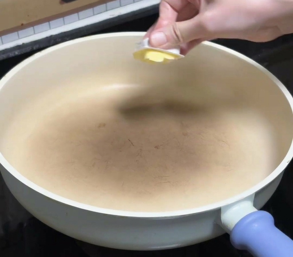 Una mano colocando un bloque cuadrado de mantequilla amarilla en una sartén cerámica blanca limpia sobre la cocina.