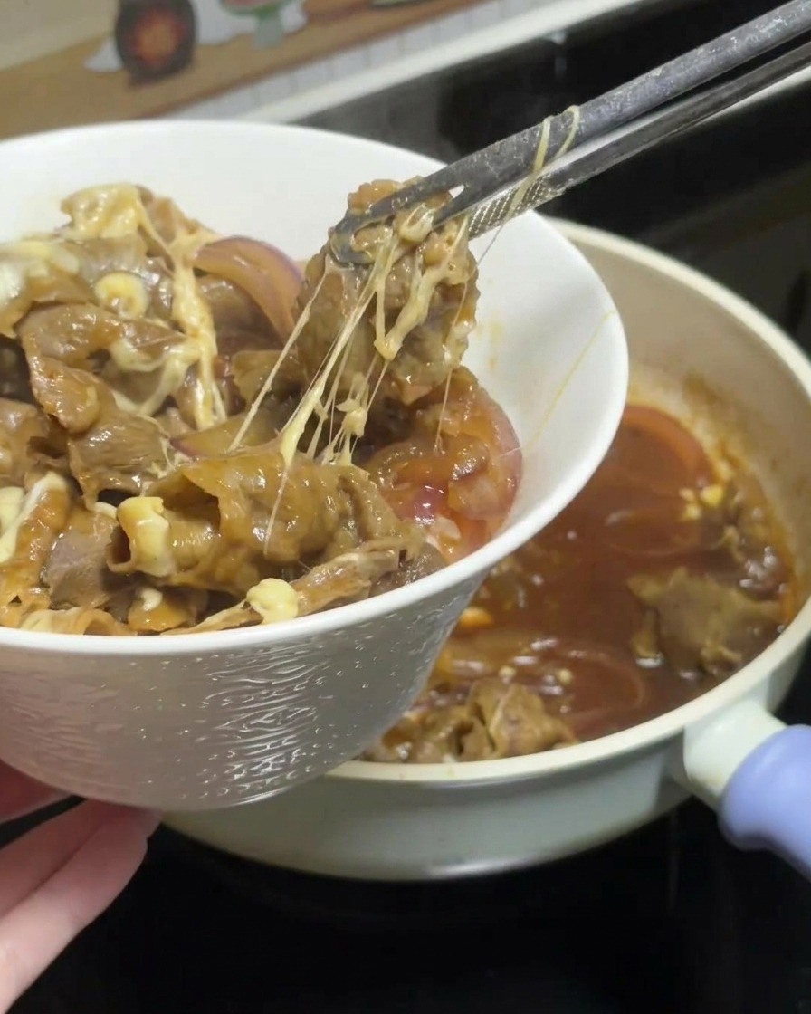 Unas pinzas levantando una porción de ternera teriyaki con queso y cebollas de la sartén, mostrando hilos largos de queso, y colocándola sobre un tazón de arroz blanco.