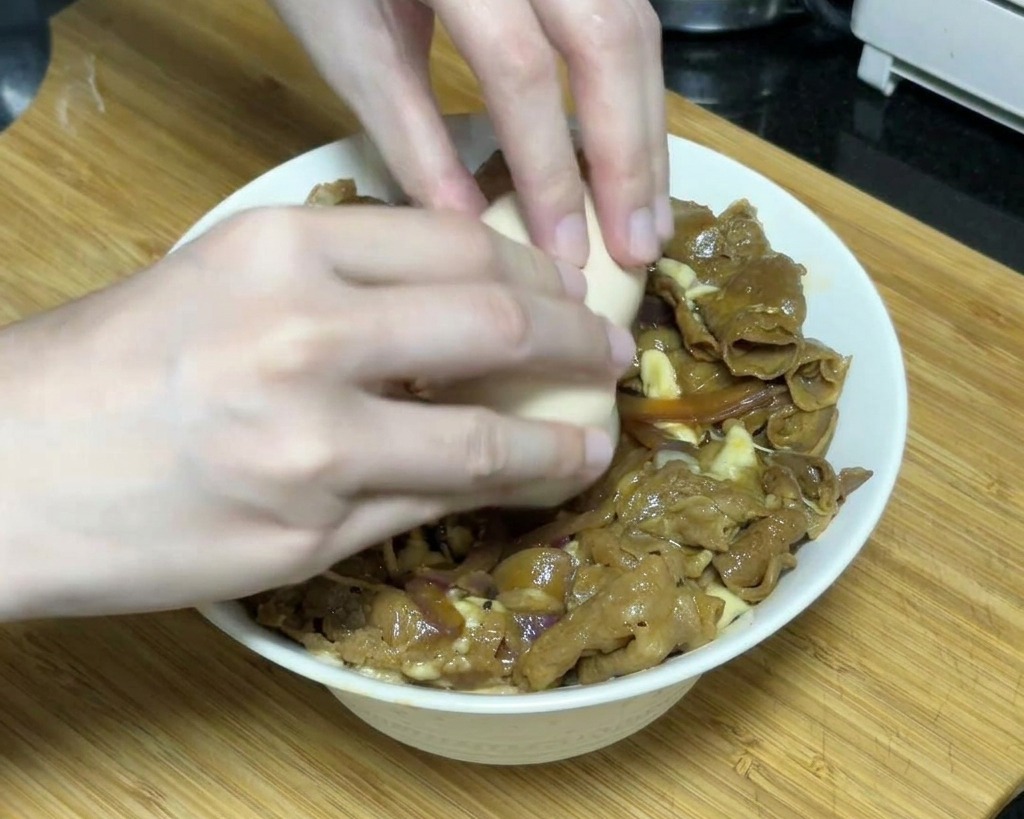 Un par de manos cascando con cuidado un huevo onsen semicocinado sobre un tazón de ternera teriyaki cubierto con queso fundido.